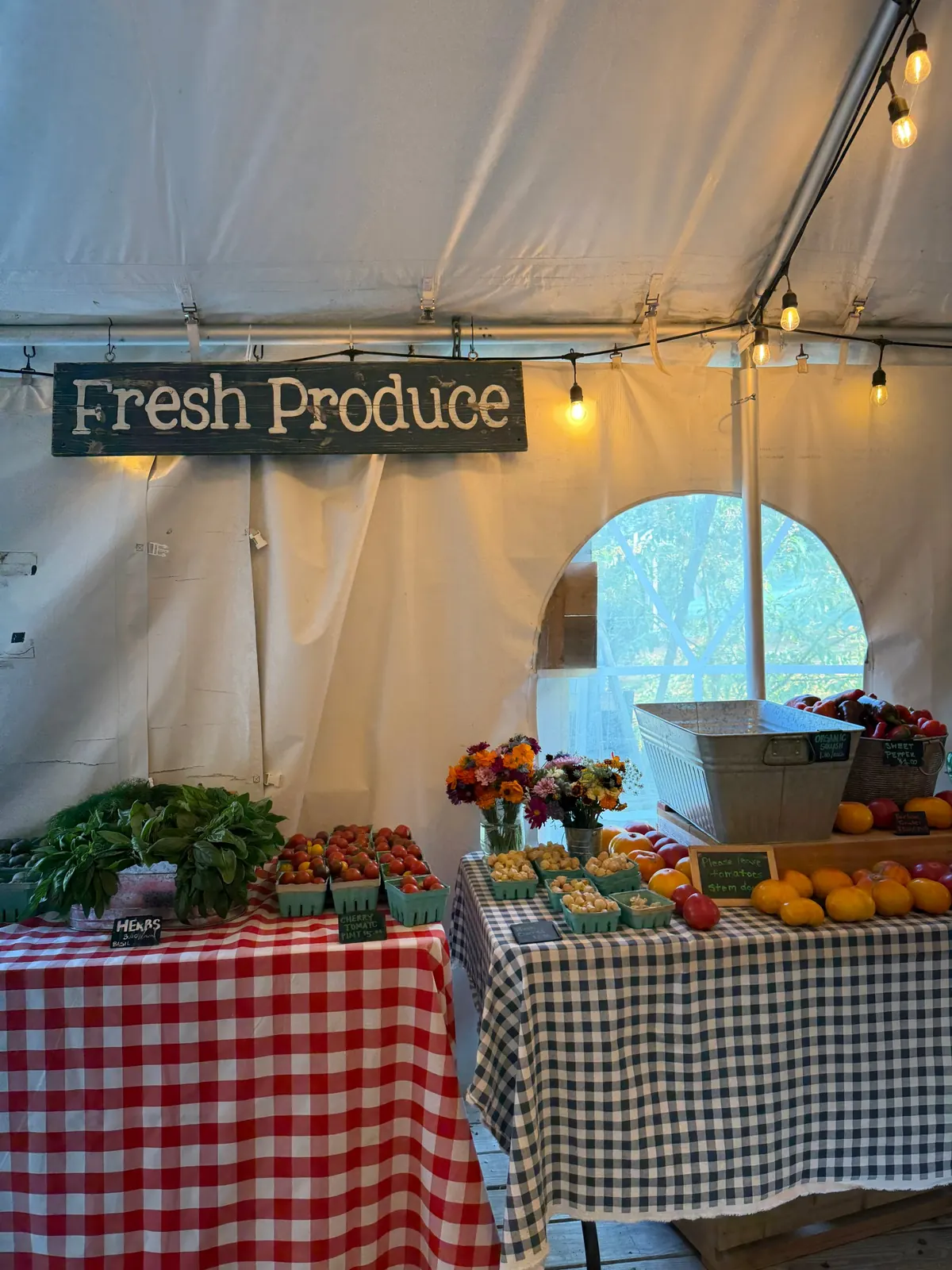 A farm stand under a tent, with a Fresh Produce sign, tomatoes, herbs and flowers on gingham cloths