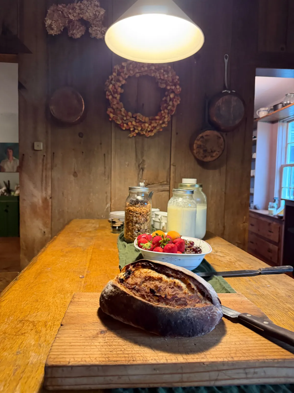 A loaf of bread on a board, with a bowl of berries and jars of milk and granola, on a long wooden farmhouse table